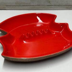 Peut inclure: Un cendrier rouge vif avec une forme unique et stylisée. Le cendrier en céramique présente une finition brillante et une série d'encoches pour reposer les cigarettes. La base a un bord marron contrastant. Un objet décoratif pour la maison.