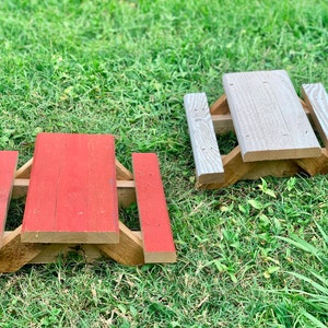 May include: Two miniature wooden picnic tables, one painted red and the other gray, are sitting on a patch of green grass.