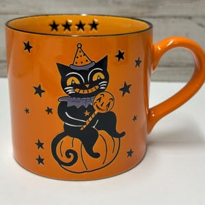 Può includere: Una tazza in ceramica arancione con l'illustrazione di un gatto nero che indossa un cappellino da festa e una gorgiera, seduto su una zucca mentre tiene un lecca-lecca. Il design include stelle nere e quattro stelle sul bordo interno.