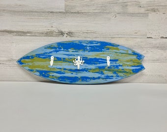 May include: A blue and green surfboard-shaped wooden wall hook with three white hooks and an octopus design.