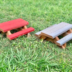 May include: Two miniature wooden picnic tables, one painted red and the other gray, sitting on a bed of green grass.