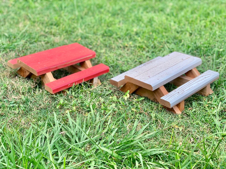 May include: Two miniature wooden picnic tables, one painted red and the other gray, sitting on a bed of green grass.