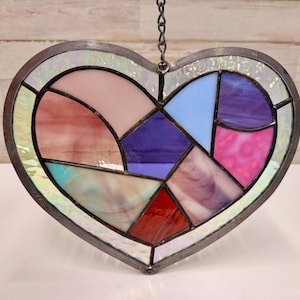 May include: A stained glass heart-shaped suncatcher with a silver-colored metal frame. The heart is divided into sections of pink, red, purple, blue, and iridescent glass. A chain is attached for hanging.