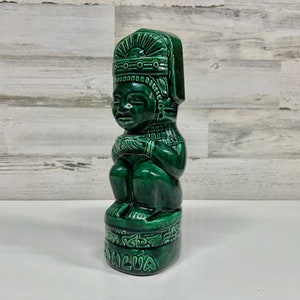 May include: Green ceramic figurine of a seated figure wearing a headdress. The figurine is decorated with intricate details and has the word "ALUA" inscribed on the base.