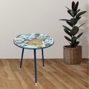 May include: A round side table with a blue metal frame and three legs. The table top is white with a printed design of bees, honeycombs, and flowers. The table is on a wood floor.