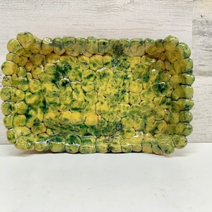 May include: A rectangular ceramic dish with a textured surface. The dish is made of small, rounded pieces of clay that are arranged in a mosaic pattern. The dish is glazed in shades of green and yellow.