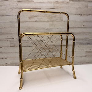 Puede incluir: Un revistero de metal dorado con un diseño estilo bambú. El revistero presenta una base rectangular, un estante de malla de alambre y un diseño de alambre entrecruzado en la parte posterior. La altura total es de aproximadamente 76 cm.