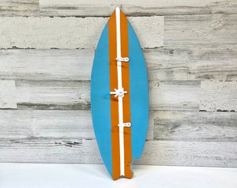 May include: A blue and orange surfboard-shaped wall hook with three white metal hooks. The surfboard is painted with a white stripe down the center.