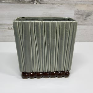 May include: A grey ceramic planter with a textured, vertical striped design. The planter has a brown, rounded base.