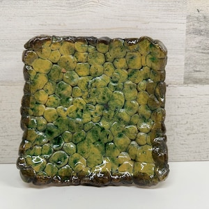 May include: A square ceramic dish with a textured surface that resembles a cobblestone path. The dish is glazed in shades of green and yellow.