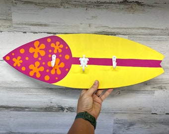 May include: A yellow surfboard-shaped wooden wall hook with a pink stripe and orange and pink floral design. The hook has three white hooks, one shaped like a turtle.