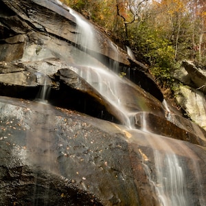 Può includere: Una cascata scenografica scende a cascata lungo una parete rocciosa scura e bagnata. L'acqua appare setosa e bianca contro la roccia scura. Il fogliame autunnale nei toni del rosso, arancione e verde incornicia la parte superiore della cascata.