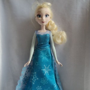 May include: A doll of a character with blonde hair in a side braid, wearing a blue dress with snowflake patterns. The dress has a sparkly bodice and a flowing skirt. The doll has blue eyes and is standing upright.