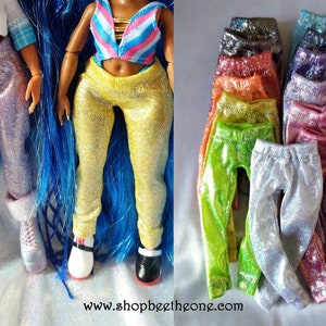 May include: Doll-sized, sparkly leggings in various colors, including silver, yellow, pink, green, and orange. Two dolls are shown wearing the leggings. The leggings are displayed in a stack on a reflective surface. The website address shopbeetheone.com is visible.