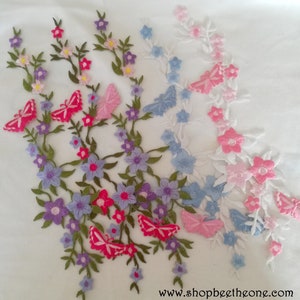 Può includere: Un tessuto bianco con ricami floreali in rosa, blu e viola. I disegni presentano farfalle e foglie.