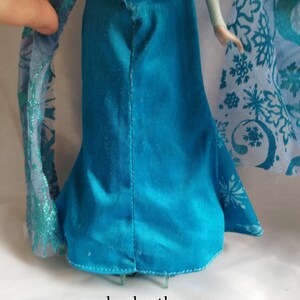 May include: A doll in a teal blue dress with a flared skirt is shown in close-up. The dress has a seam down the back. A sheer, light blue shawl with snowflake patterns is visible. The website address www.shopbeethetone.com is at the bottom.