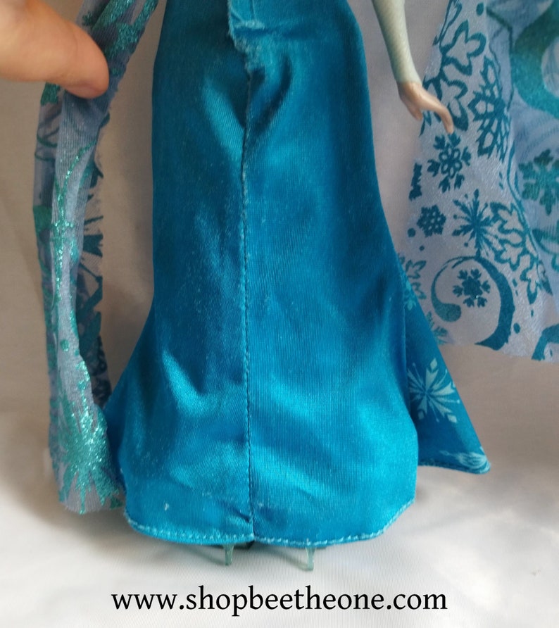 May include: A doll in a teal blue dress with a flared skirt is shown in close-up. The dress has a seam down the back. A sheer, light blue shawl with snowflake patterns is visible. The website address www.shopbeethetone.com is at the bottom.