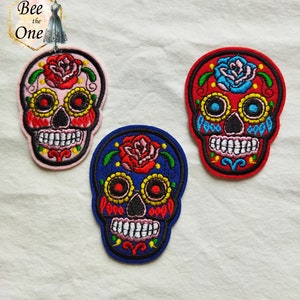 Può includere: Tre toppe ricamate di teschio di zucchero in rosa, rosso e blu. Ogni teschio ha una rosa rossa, occhi neri e denti bianchi.