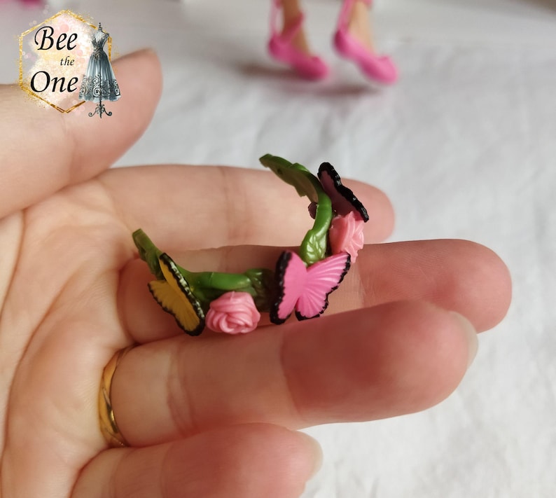 以下が含まれることがあります： ピンクの花と蝶がついた緑色のプラスチック製のヘッドバンド。ヘッドバンドはピンクと黄色の蝶とピンクの花で飾られています。