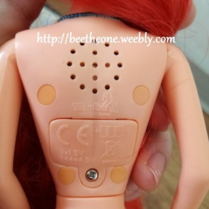 May include: A red-haired doll with a light pink body and a blue top. The doll has a speaker on its back with a small speaker grill and a label that says "Simba Made in China". The doll also has a battery compartment with the text "3x1.5V R44=4.5V" and a CE mark.