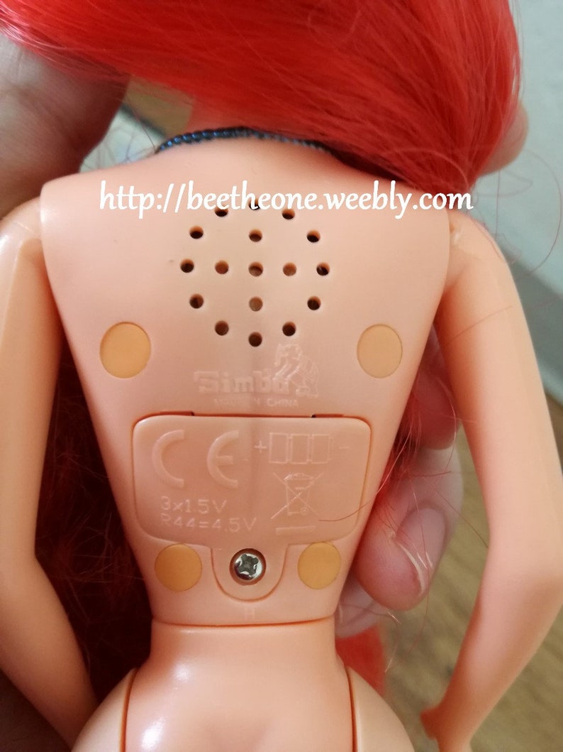 May include: A red-haired doll with a light pink body and a blue top. The doll has a speaker on its back with a small speaker grill and a label that says "Simba Made in China". The doll also has a battery compartment with the text "3x1.5V R44=4.5V" and a CE mark.