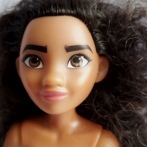 Pode incluir: Um close-up do rosto de uma boneca com olhos castanhos, cabelo castanho escuro e tez marrom claro. A boneca est&aacute; usando batom rosa e tem uma express&atilde;o neutra.
