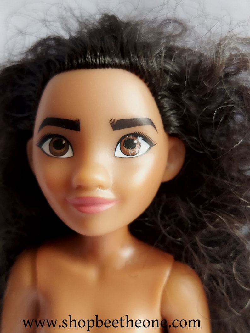 Pode incluir: Um close-up do rosto de uma boneca com olhos castanhos, cabelo castanho escuro e tez marrom claro. A boneca est&aacute; usando batom rosa e tem uma express&atilde;o neutra.