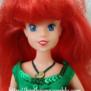 May include: A red-haired doll with green eyes wearing a green sequined dress and a necklace with a seashell pendant. The doll's hair is styled in a long, flowing mane. The doll's eyes are large and expressive. The dress is adorned with sequins that shimmer in the light. The necklace is a simple chain with a seashell pendant that hangs in the center of the doll's chest. The doll is a popular character from the Disney movie The Little Mermaid.