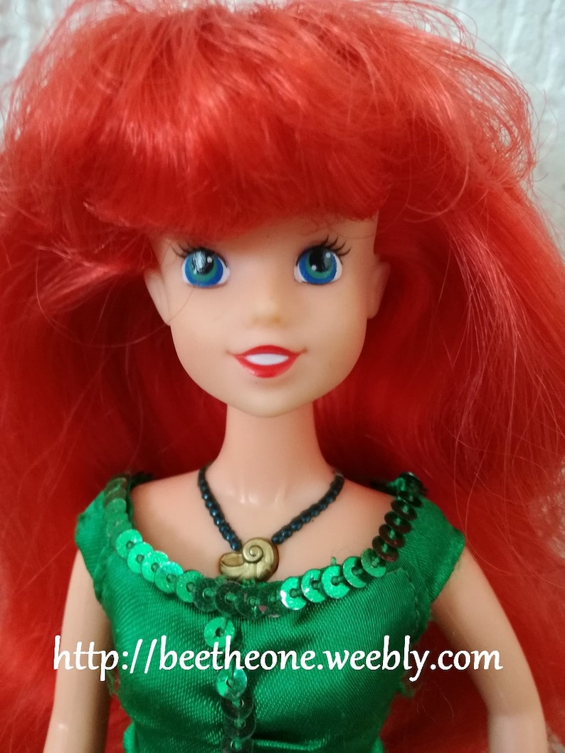 May include: A red-haired doll with green eyes wearing a green sequined dress and a necklace with a seashell pendant. The doll's hair is styled in a long, flowing mane. The doll's eyes are large and expressive. The dress is adorned with sequins that shimmer in the light. The necklace is a simple chain with a seashell pendant that hangs in the center of the doll's chest. The doll is a popular character from the Disney movie The Little Mermaid.