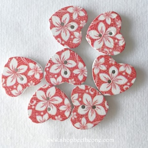 Puede incluir: Seis botones rojos en forma de corazón con diseños florales blancos. Los botones tienen dos agujeros cada uno.