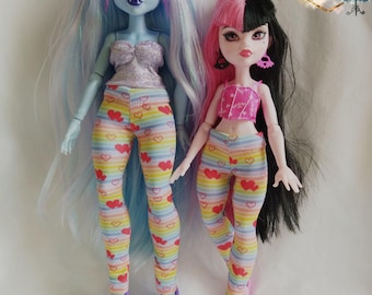 Könnte beinhalten: Zwei Modepuppen tragen Regenbogen-Leggings mit einem Herzmuster. Die Puppe links hat blaues Haar und trägt ein silbernes Oberteil. Die Puppe rechts hat schwarz-pinkes Haar und trägt ein pinkes Oberteil.  "Bee the One" ist in goldenen Buchstaben auf weißem Hintergrund geschrieben.
