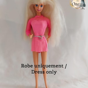 Può includere: Una bambola con lunghi capelli biondi e occhi blu che indossa un vestito rosa con scollatura a rete e maniche lunghe. L'abito è stretto in vita con una cintura argentata. Il testo "Robe uniquement / Dress only" è sotto.