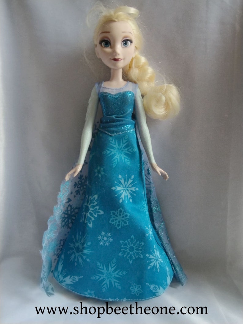 May include: A doll of a character with blonde hair in a side braid, wearing a blue dress with snowflake patterns. The dress has a sparkly bodice and a flowing skirt. The doll has blue eyes and is standing upright.