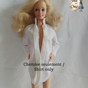 Peut inclure: Une poupée porte une chemise blanche à col large et à manches longues. La chemise est ouverte sur le devant. Le texte "Chemise seulement / Shirt only" est imprimé sur l'image.