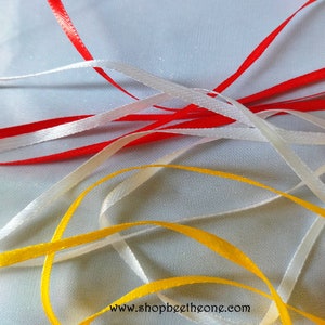 Peut inclure: Gros plan sur quatre rubans de satin de couleur rouge, blanc et jaune. Les rubans sont disposés en croisillon sur une surface blanche. Le texte "www.shopbeettheone.com" est visible en bas de l'image.