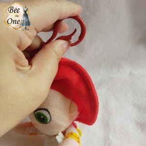 K&ouml;nnte beinhalten: Ein rotes Pl&uuml;schtier mit einem roten Hut und einem gr&uuml;nen Auge. Das Spielzeug hat einen roten Plastikclip, der an der Oberseite des Kopfes befestigt ist.