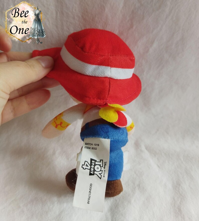 K&ouml;nnte beinhalten: Rotes und wei&szlig;es Pl&uuml;schtier von Woody aus Toy Story 4. Woody tr&auml;gt seinen charakteristischen Cowboyhut und seine Weste. Das Spielzeug ist etwa 15 cm gro&szlig;. Das Etikett auf dem Spielzeug lautet "Disney Pixar Toy Story 4".