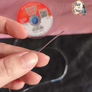 Op de afbeelding: Een helder, flexibel elastisch sieraadkoord voor kralenprojecten. Het koord is dun en transparant en wordt getoond terwijl het uit een ronde spoel komt met het label Jewelry Elastic Cord, een blauw logo en een QR-code op de achtergrond.