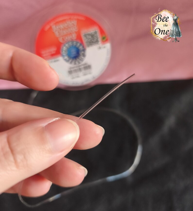 Op de afbeelding: Een helder, flexibel elastisch sieraadkoord voor kralenprojecten. Het koord is dun en transparant en wordt getoond terwijl het uit een ronde spoel komt met het label Jewelry Elastic Cord, een blauw logo en een QR-code op de achtergrond.
