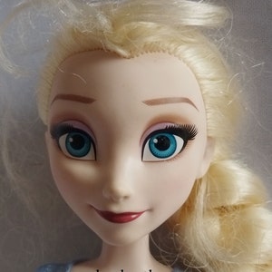 May include: A close-up of a doll's face with blonde hair and blue eyes. The doll has long eyelashes, pink eyeshadow, and a red lipstick smile. The doll's face is pale with rosy cheeks. The website address www.shopbeetheone.com is visible at the bottom.