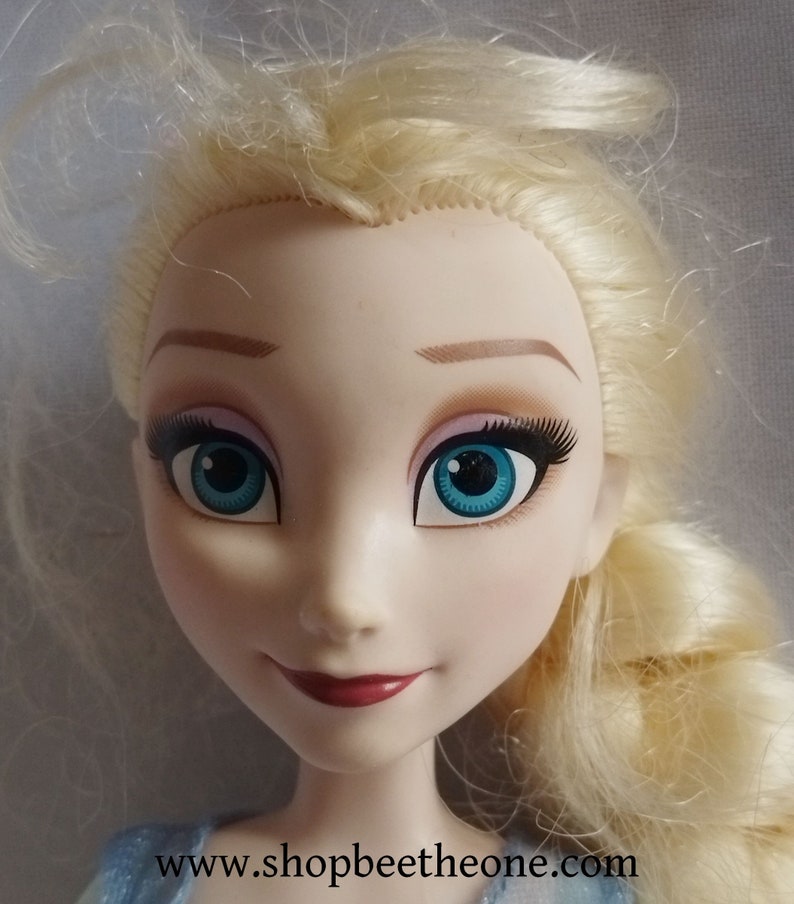 May include: A close-up of a doll's face with blonde hair and blue eyes. The doll has long eyelashes, pink eyeshadow, and a red lipstick smile. The doll's face is pale with rosy cheeks. The website address www.shopbeetheone.com is visible at the bottom.