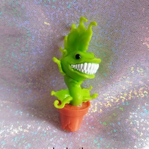 Peut inclure: Une plante en plastique verte et blanche avec un sourire narquois. La plante est dans un petit pot orange.