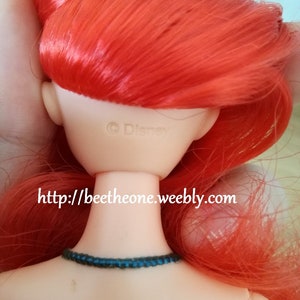 May include: A red-haired doll with a blue necklace. The doll's back is visible, showing the text "&copy; Disney" and a website address.