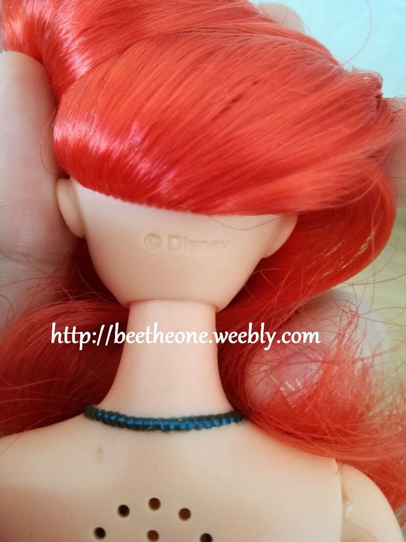 May include: A red-haired doll with a blue necklace. The doll's back is visible, showing the text "&copy; Disney" and a website address.