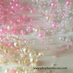 Peut inclure: Une guirlande de perles blanches et roses enfilées sur un fil transparent. La guirlande est composée de petites et de grandes perles.