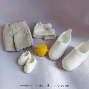 May include: A set of doll clothes including a pair of white trainers, a pair of smaller white trainers, a pair of khaki shorts, a white shirt, and a yellow plastic container.