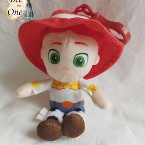K&ouml;nnte beinhalten: Ein Pl&uuml;schtier von Woody aus Toy Story, das seinen charakteristischen roten Hut und sein Cowboy-Outfit tr&auml;gt. Das Spielzeug hat einen Clip auf der R&uuml;ckseite, wodurch es zu einem Schl&uuml;sselanh&auml;nger oder einem Taschenanh&auml;nger wird.
