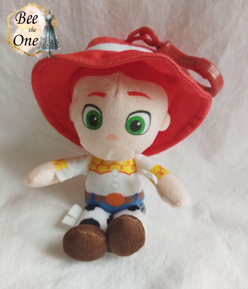 K&ouml;nnte beinhalten: Ein Pl&uuml;schtier von Woody aus Toy Story, das seinen charakteristischen roten Hut und sein Cowboy-Outfit tr&auml;gt. Das Spielzeug hat einen Clip auf der R&uuml;ckseite, wodurch es zu einem Schl&uuml;sselanh&auml;nger oder einem Taschenanh&auml;nger wird.
