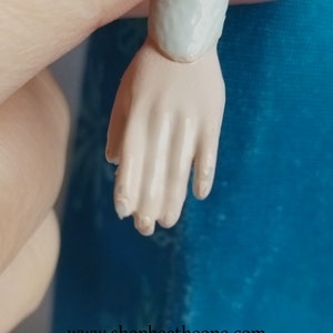 May include: Close-up of a doll's hand with a light skin tone. The fingers are slightly bent, and the nails are visible. The doll's arm is attached to the hand. The background is a solid blue colour. The website address www.shopbeetheone.com is visible at the bottom.