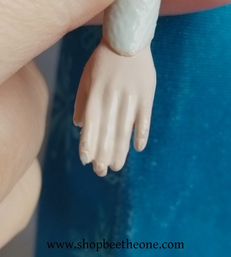 May include: Close-up of a doll's hand with a light skin tone. The fingers are slightly bent, and the nails are visible. The doll's arm is attached to the hand. The background is a solid blue colour. The website address www.shopbeetheone.com is visible at the bottom.